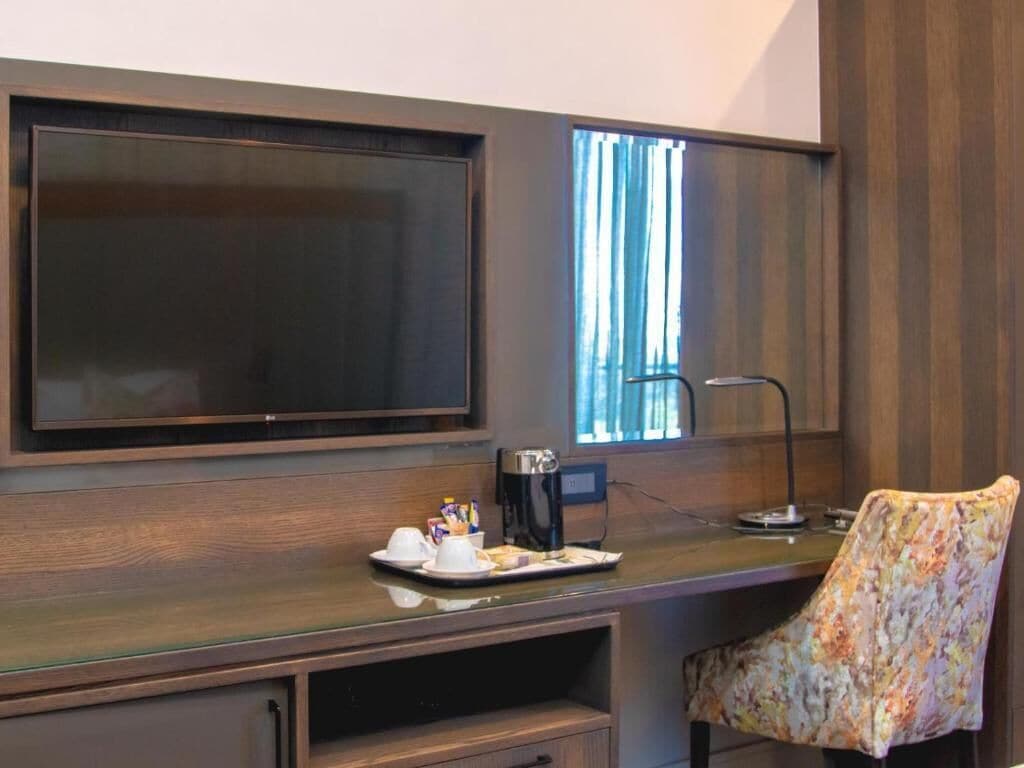 A room with a large flat-screen TV mounted on the wall, a desk with a coffee maker, a chair, and a window with curtains.
