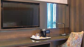 A room with a large flat-screen TV mounted on the wall, a desk with a coffee maker, a chair, and a window with curtains.