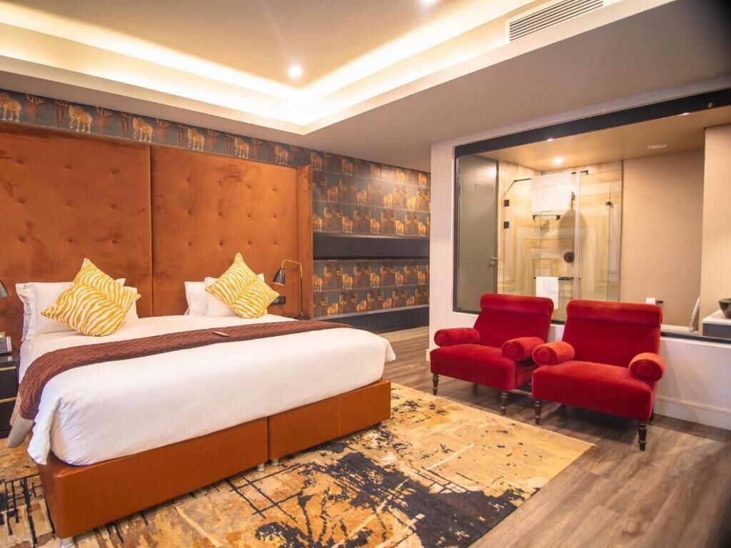A modern hotel room with a large bed, red chairs, a patterned rug, and a decorative wall.