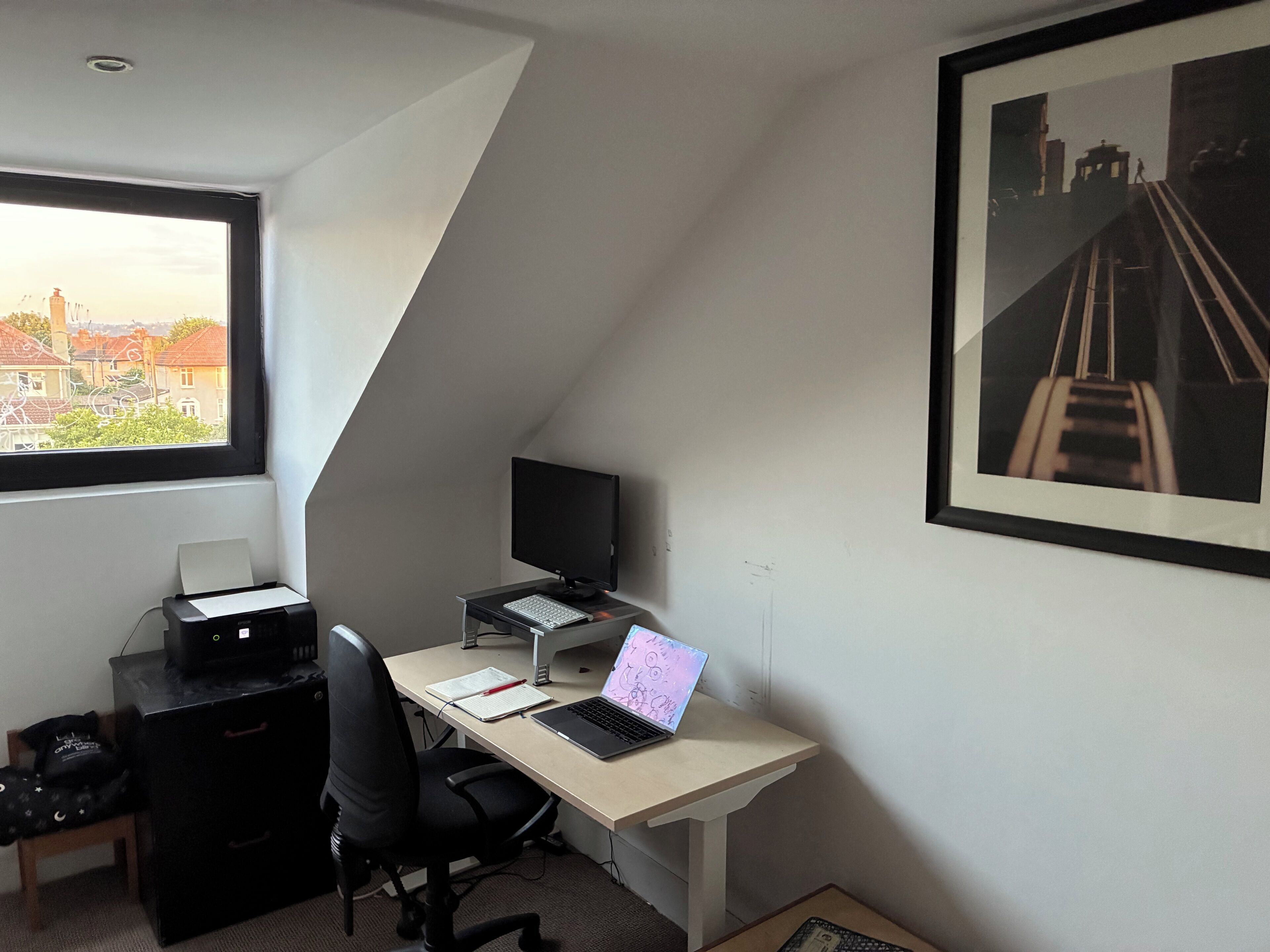 Een kamer met een bureau, computer, stoel, printer en een raam met uitzicht op de stad.