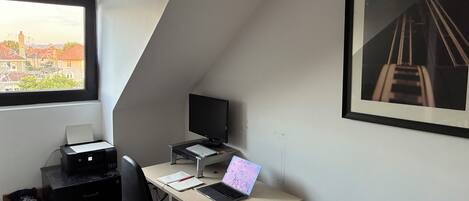 Een kamer met een bureau, computer, stoel, printer en een raam met uitzicht op de stad.