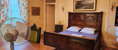 A bedroom with a wooden bed, a standing fan, a chandelier, a painting, and a door.