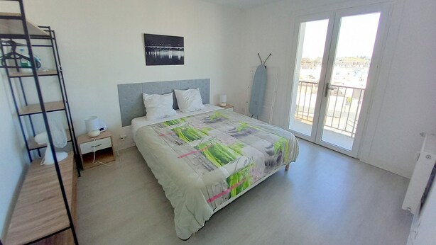Een slaapkamer met een bed, nachtkastje, plank, plafondventilator, een foto aan de muur en een balkon.