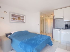 Ein Schlafzimmer mit Bett, Nachttisch, kleinem Schreibtisch, Stuhl, Kleiderschrank und Spiegel, sowie einer Küchenzeile mit Kühlschrank und Kaffeemaschine.