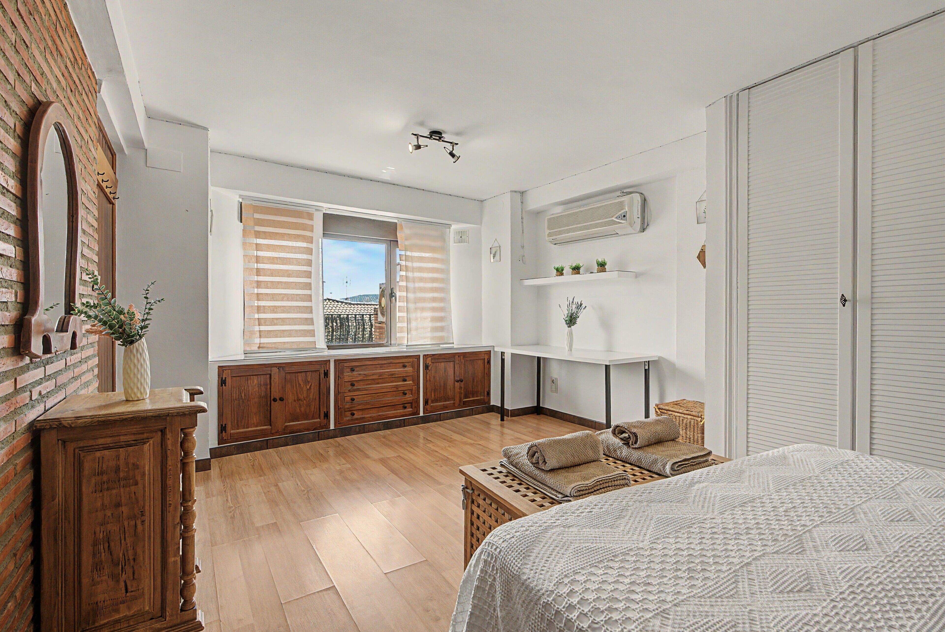 Un dormitorio con cama, armario de madera, ventana con persianas, unidad de aire acondicionado blanca y una mesita con toallas.