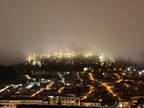 Un paisaje urbano histórico con una fortaleza imponente en la cima de una colina, calles serpenteantes y edificios iluminados.