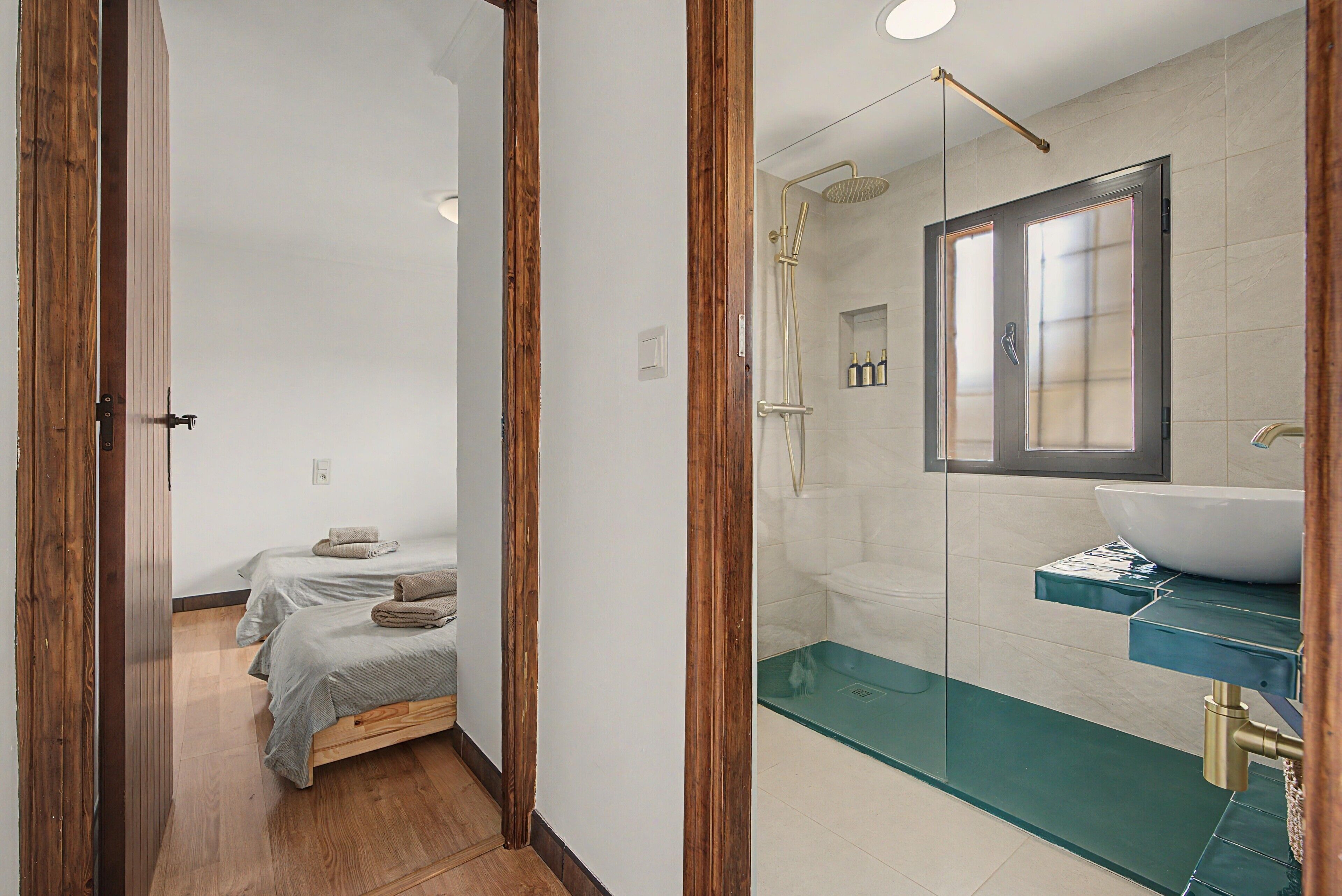 A modern bathroom with a glass shower enclosure, a sink with a blue countertop, and a bed with neatly folded towels.