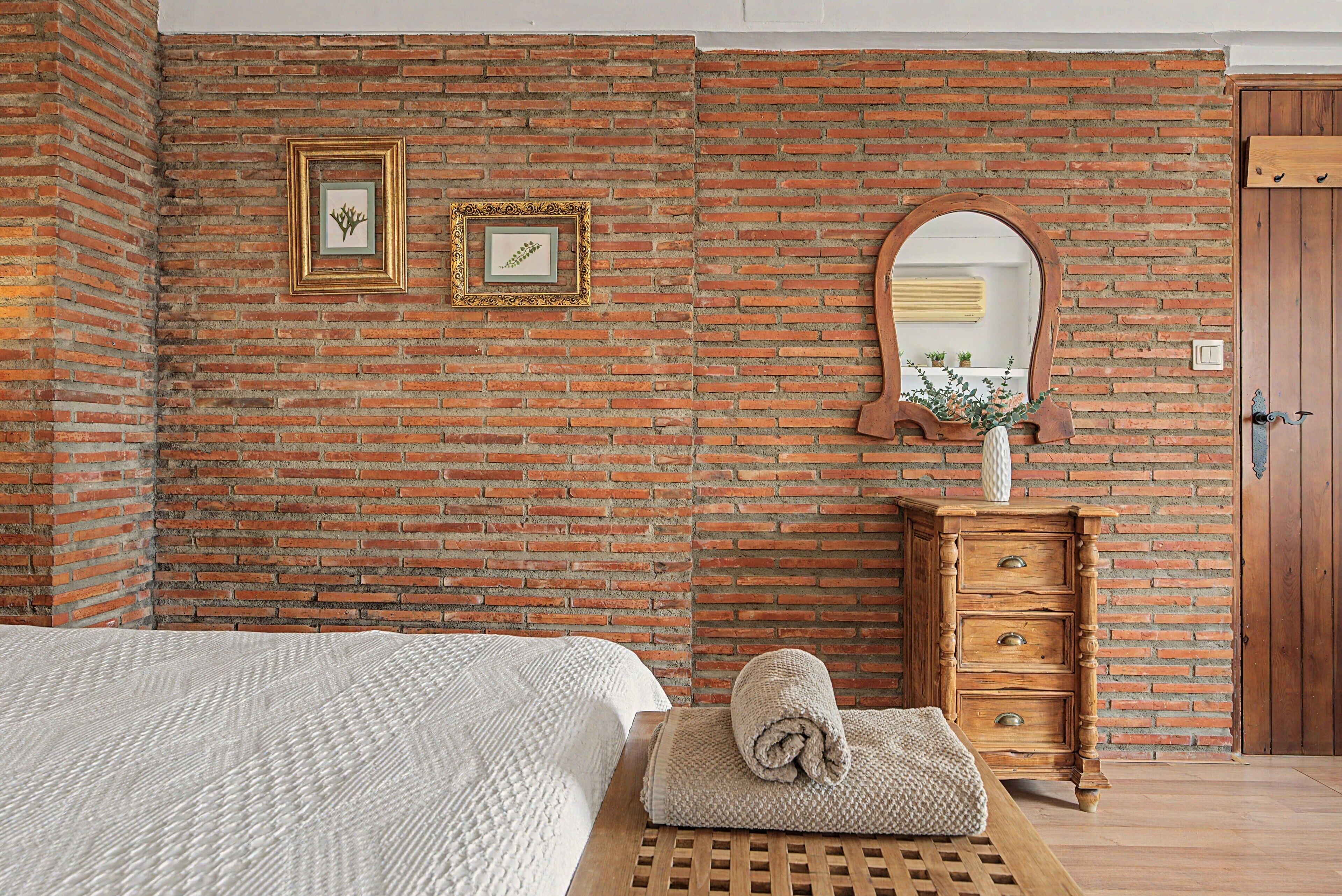 Una habitación con pared de ladrillo, una cama con manta blanca, un aparador de madera y un espejo.