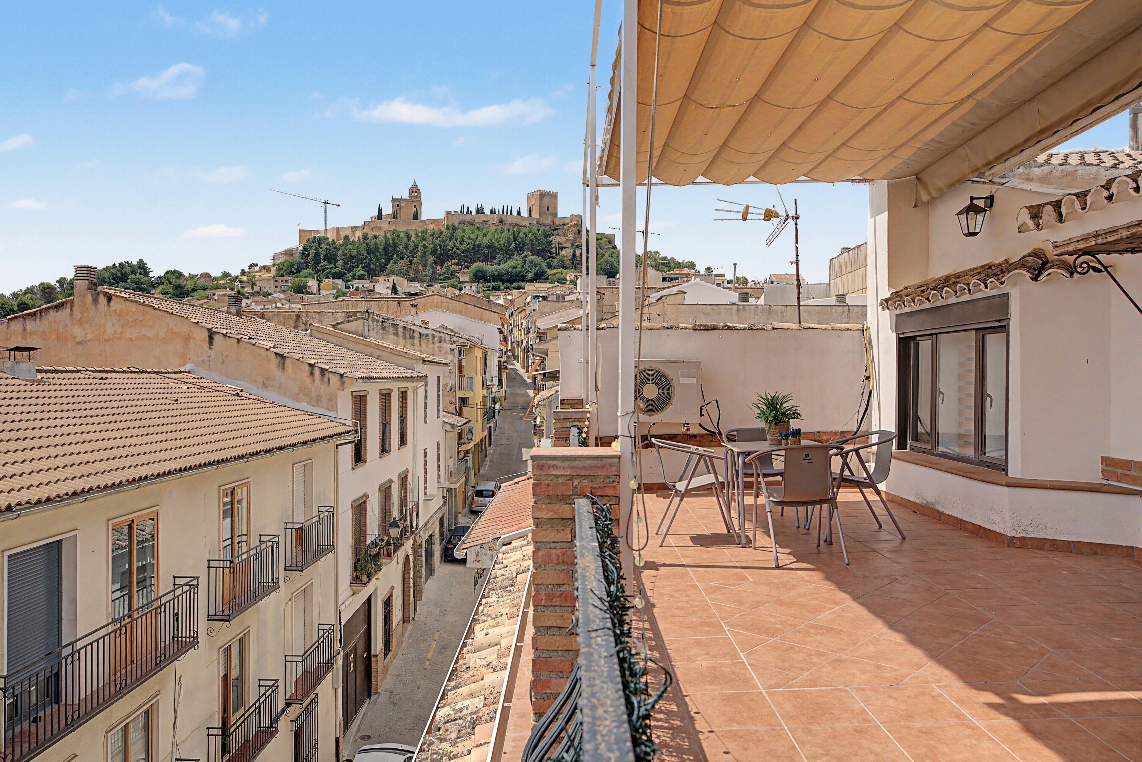 Una terraza con vistas a un castillo histórico en una colina, tejado de baldosas y una mesa con sillas.