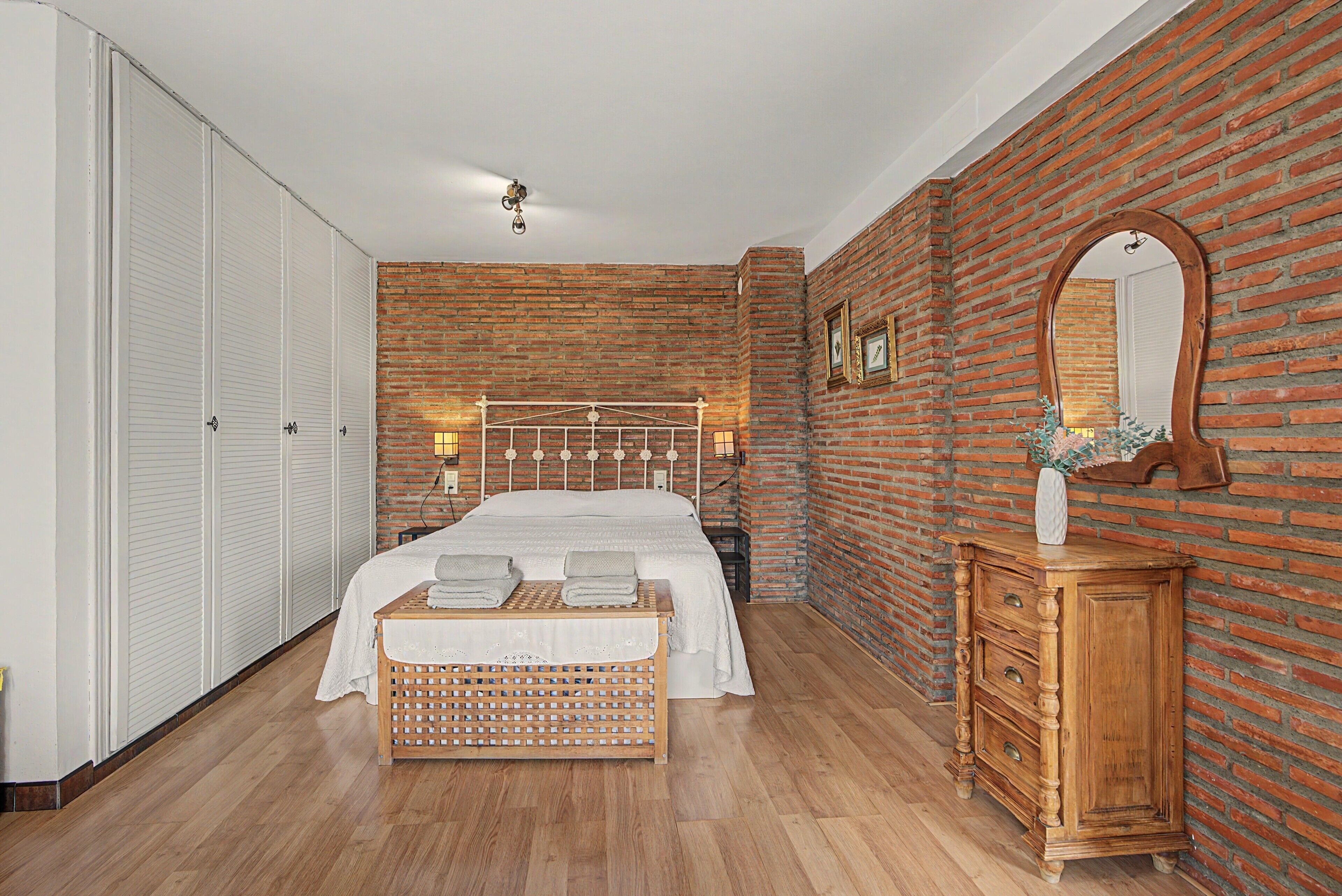 Une chambre avec un mur en briques, un cadre de lit en bois, une commode en bois et une armoire blanche.