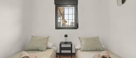 Una habitación pequeña y limpia con dos camas individuales, una mesita, una ventana con vistas y un espejo colgado.