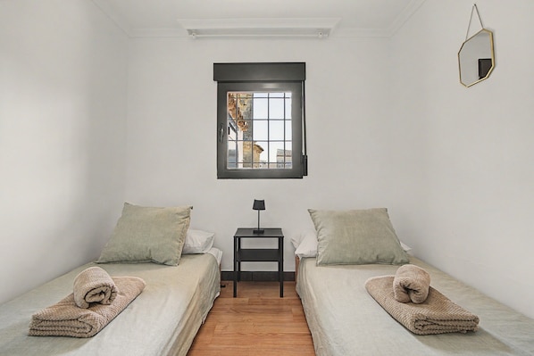 A small, clean room with two single beds, a small table, a window with a view, and a hanging mirror.