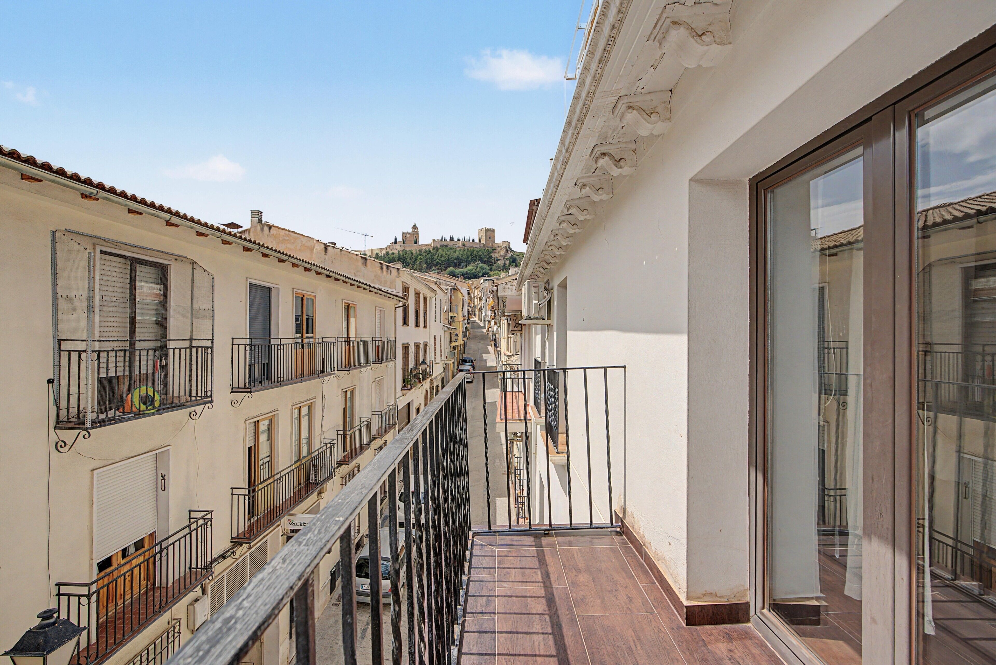 Une rue étroite bordée de bâtiments des deux côtés, un balcon avec une balustrade en métal, et une vue sur une colline où l’on aperçoit, au loin, une structure ressemblant à un château.