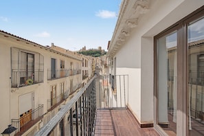 A narrow street with buildings on either side, a balcony with a metal railing, and a view of a hill with a castle-like structure in the distance.
