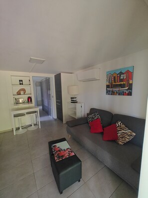 A living room with a grey sofa, a black ottoman, a white table with two stools, a painting on the wall, and a kitchen area in the background. - Near Bordeaux: air conditioning, parking, and a picturesque setting (Latresne)