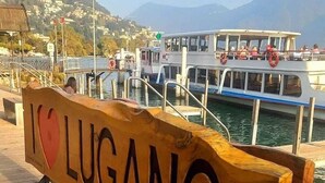 Et træskilt med teksten 'I LOVE LUGANO', en dok, en båd og et bjergrigt landskab i baggrunden.