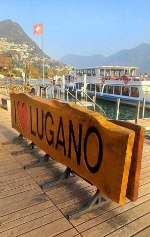 Un panneau en bois sur lequel on peut lire « J’AIME LUGANO », un quai, un bateau et, au loin, un paysage montagneux.