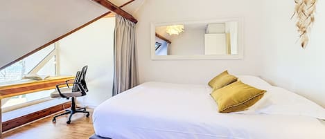 A bedroom with a large bed, a desk with a chair, a window, and a mirror on the wall.