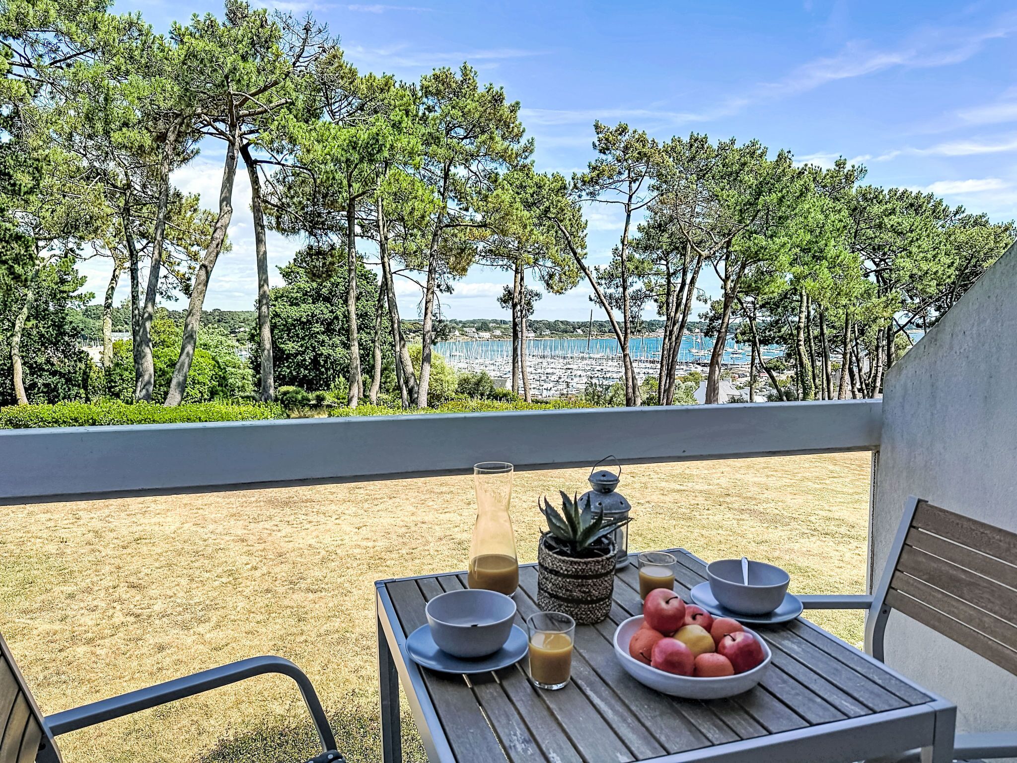 A table with bowls, apples, and drinks on a balcony overlooking a beach with trees and boats.