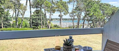 Een tafel met kommen, appels en drankjes op een balkon met uitzicht op een strand met bomen en boten.