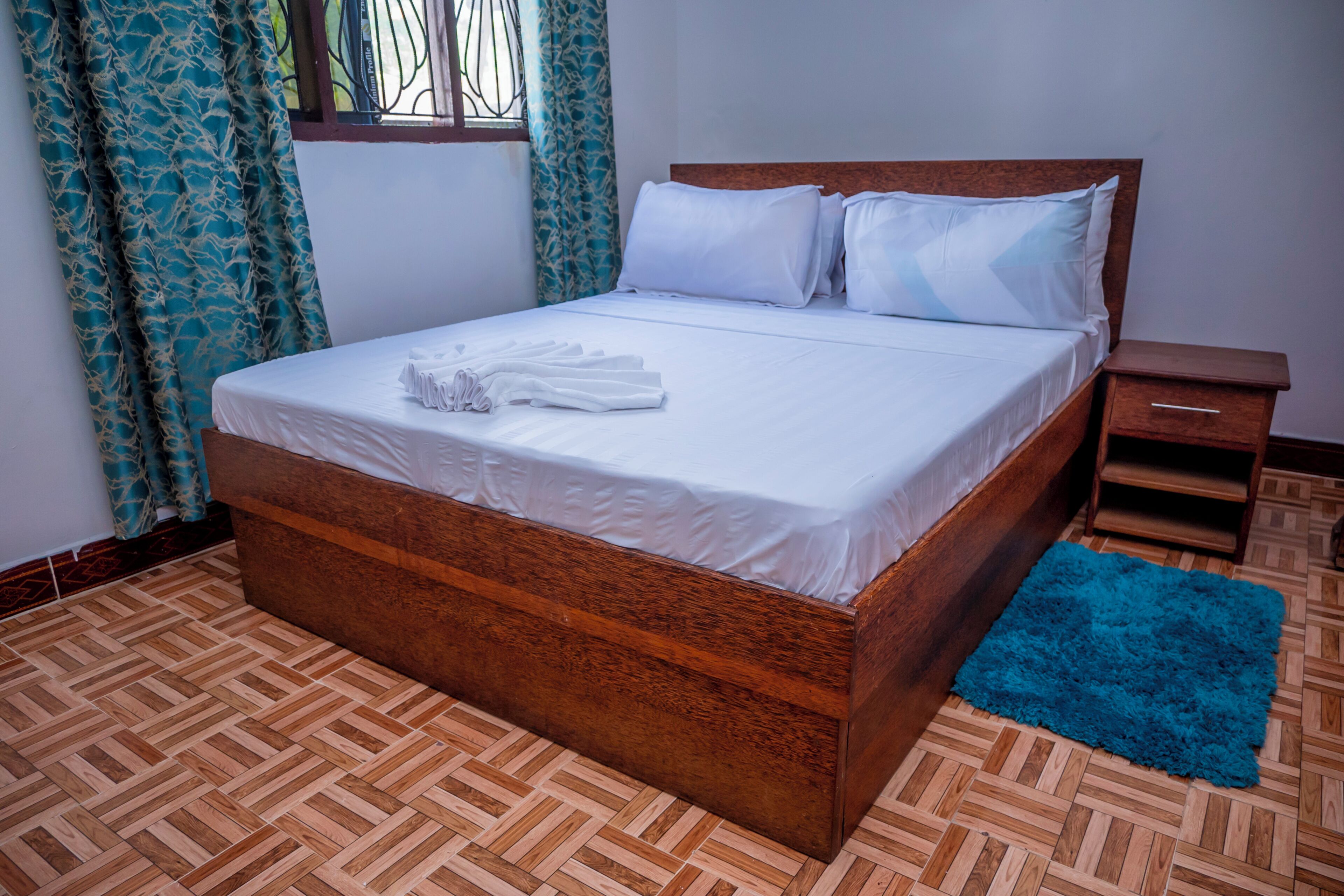 Un lit bien fait, avec du linge de lit blanc, une tête de lit en bois et un tapis bleu sur un plancher en parquet.