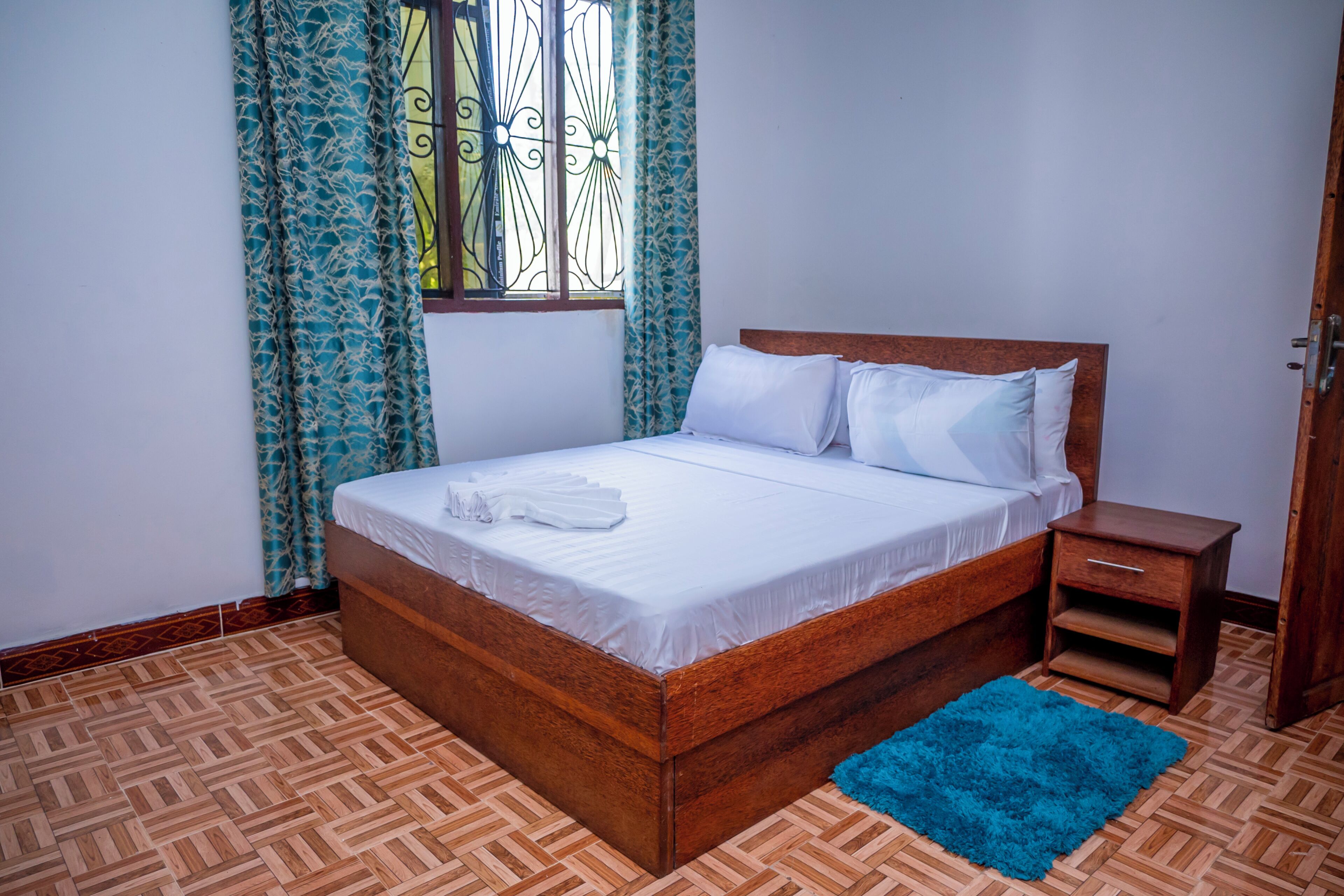 A bedroom with a wooden bed, a nightstand, a blue rug, and patterned curtains.