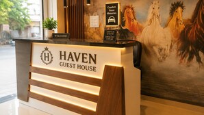 A reception desk with 'HAVEN GUEST HOUSE' signage, a 'WELCOME' sign, a 'CHECK-IN 12:00 PM CHECK-OUT 11:00 AM' sign, a 'Comfort Peace Memories' sign, and a painting of horses.