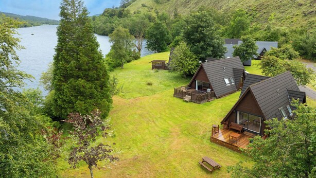 Eine Blockhütte am Seeufer mit einer Terrasse, umgeben von üppiger Vegetation und Hügeln.