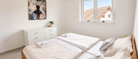 A bedroom with two beds, a dresser, a window with a view of a house, and a painting on the wall.