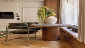 Una habitación moderna con una mesa de comedor de madera, dos sillas tapizadas de verde, un jarrón blanco grande con una planta colgante, un banco empotrado con cojines, un televisor de pantalla plana montado en la pared y una ventana con cortinas blancas.