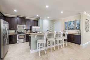A modern kitchen with dark cabinets, stainless steel appliances, a central island with bar stools, and decorative wall art. - Arcade Games Room + Private Pool | Mins to Disney | Cosmic Clubhouse (Kissimmee)