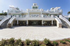 Um edifício grande, com vários andares, entrada central, diversas varandas e uma torre com relógio.