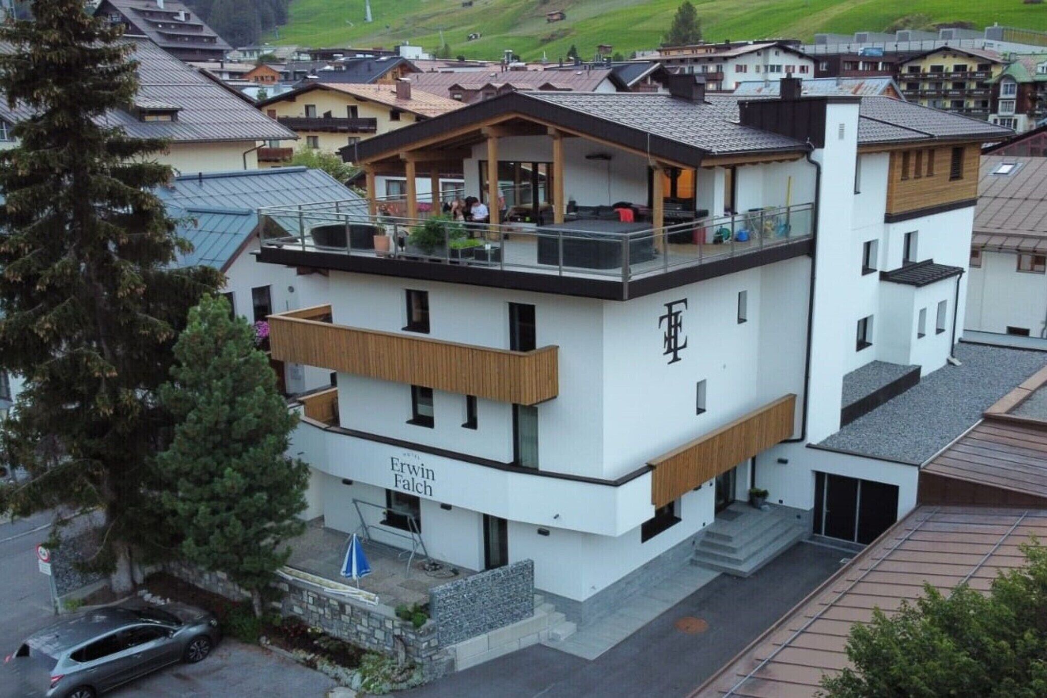 A multi-story building with a balcony, a sign reading 'Erwin Falch,' and a view of a residential area with houses and a hill in the background.