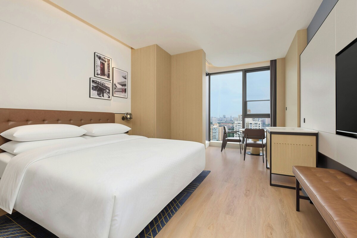 A hotel room with a large bed, a desk, a chair, and a view of the city through the window.