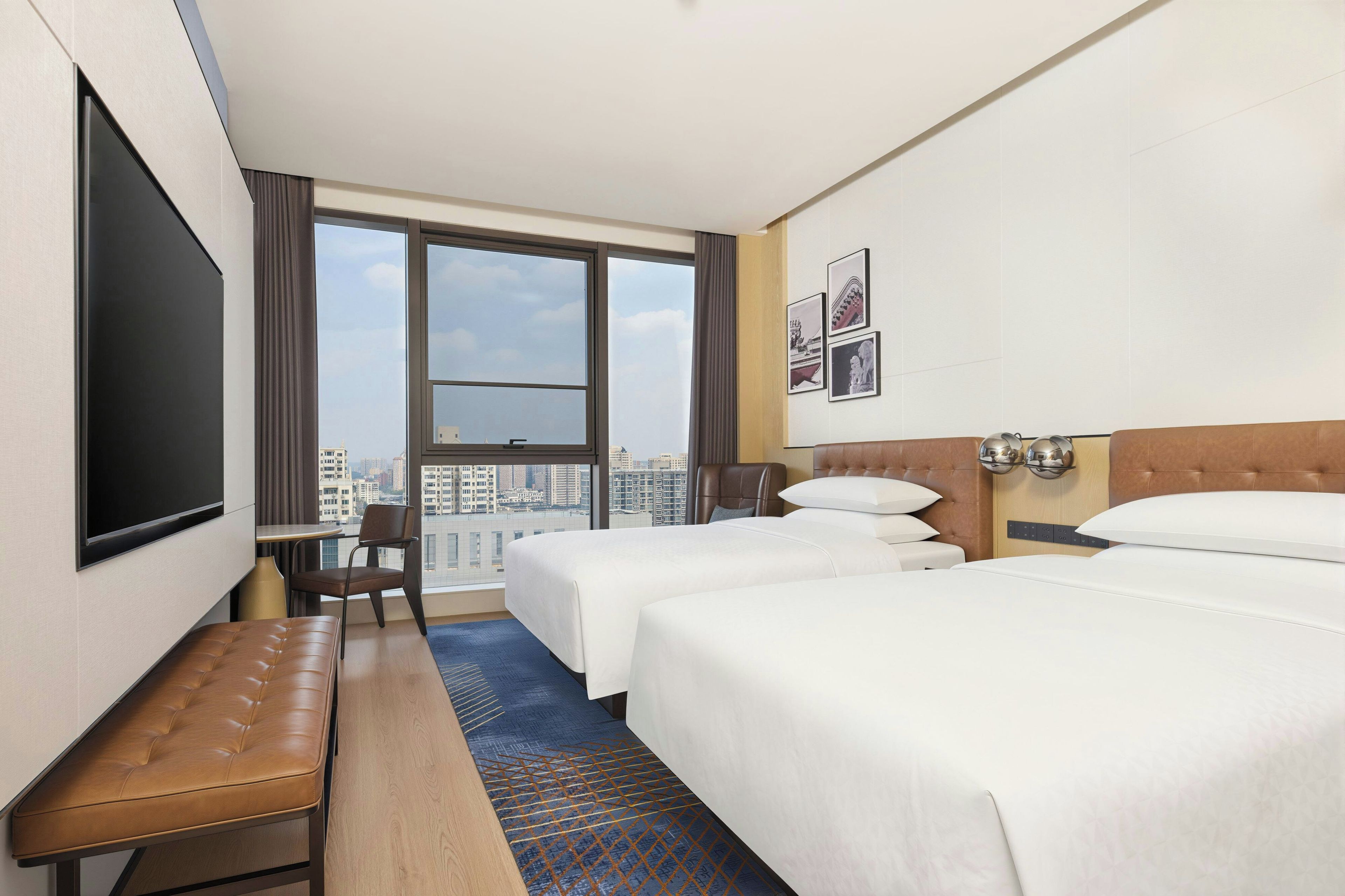 A hotel room with two beds, a desk, a chair, a bench, and a large window offering a city view.