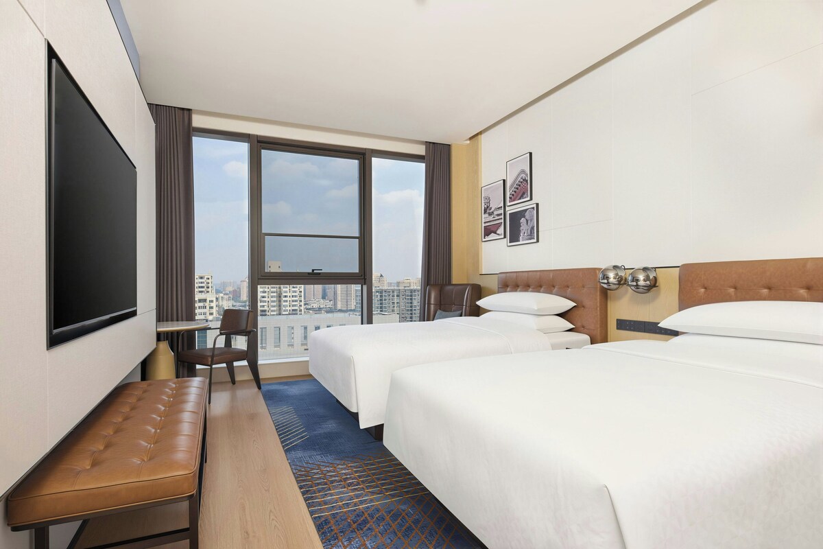 A hotel room with two beds, a desk, a chair, a bench, and a large window offering a city view.