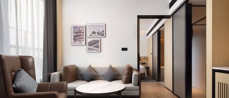 Habitación de hotel moderna con sofá, sillón y una mesita. Hay cuadros en la pared, un ventanal con cortinas y una puerta que da a otra habitación.