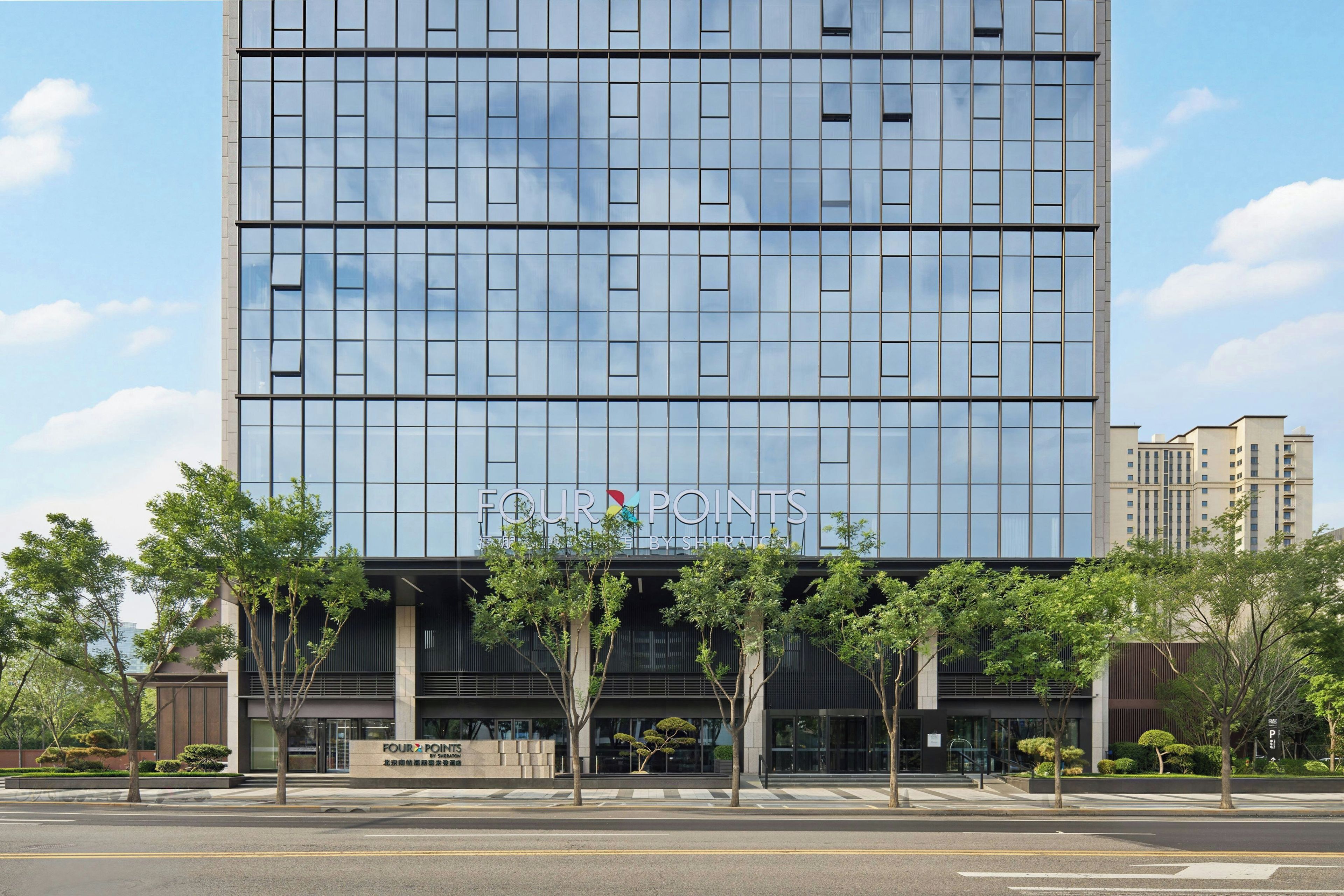 Un bâtiment moderne doté d’une façade en verre, d’une enseigne « FOUR POINTS » bien visible et d’une entrée aménagée avec des arbres.