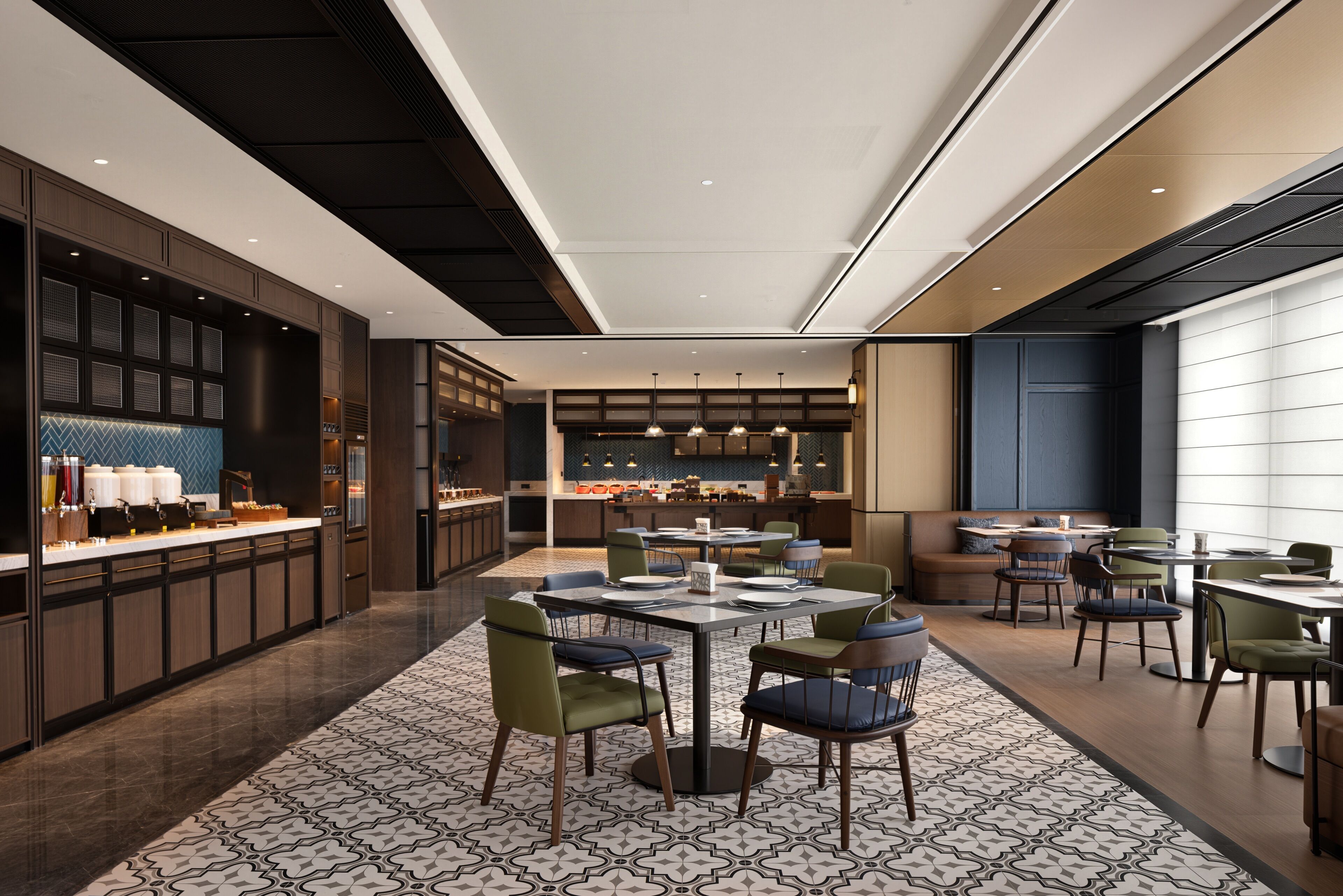 A modern hotel lobby with a central dining area, a bar, and a kitchen visible in the background.