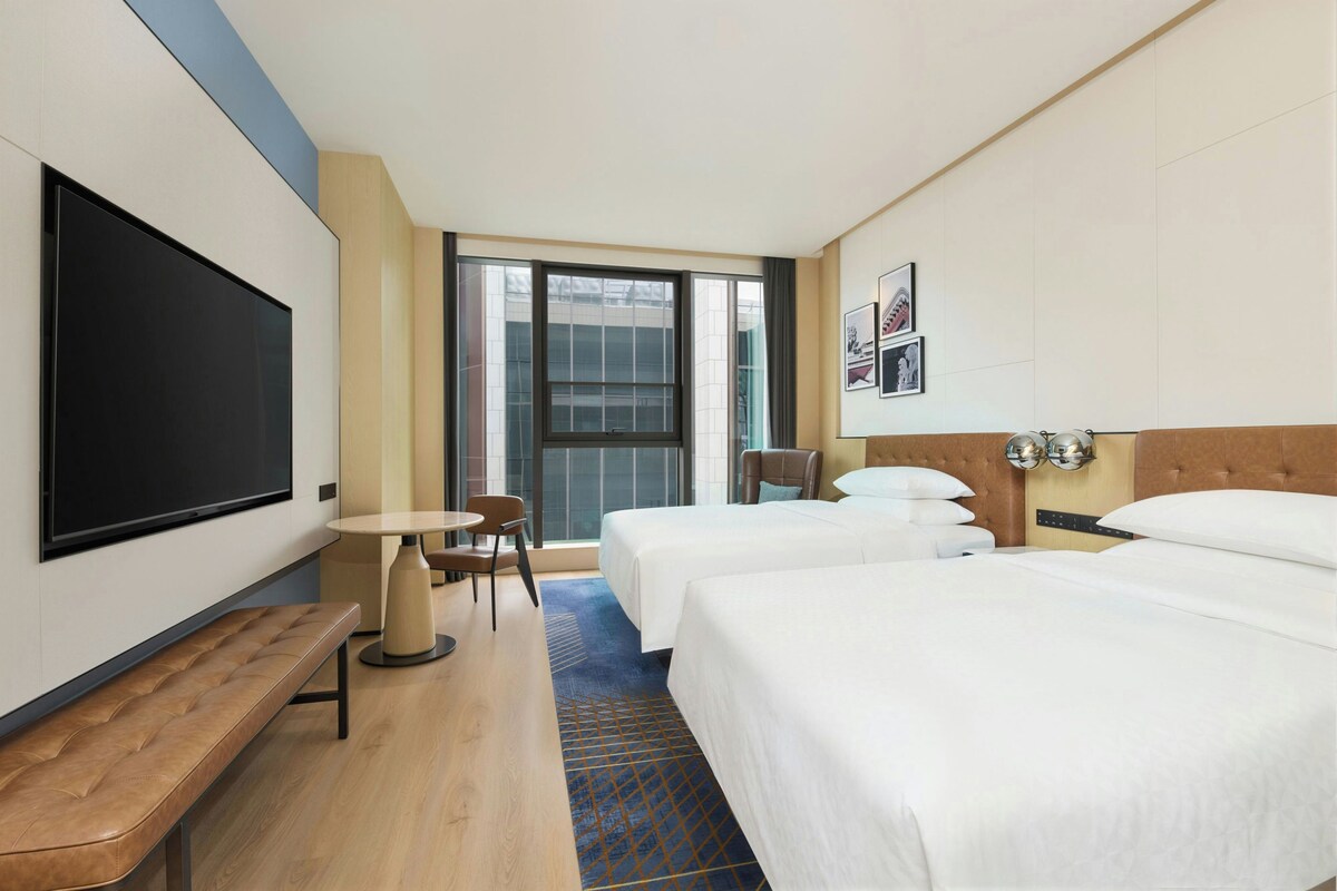 A hotel room with two beds, a flat-screen TV, a small table, a bench, and a window with a view.