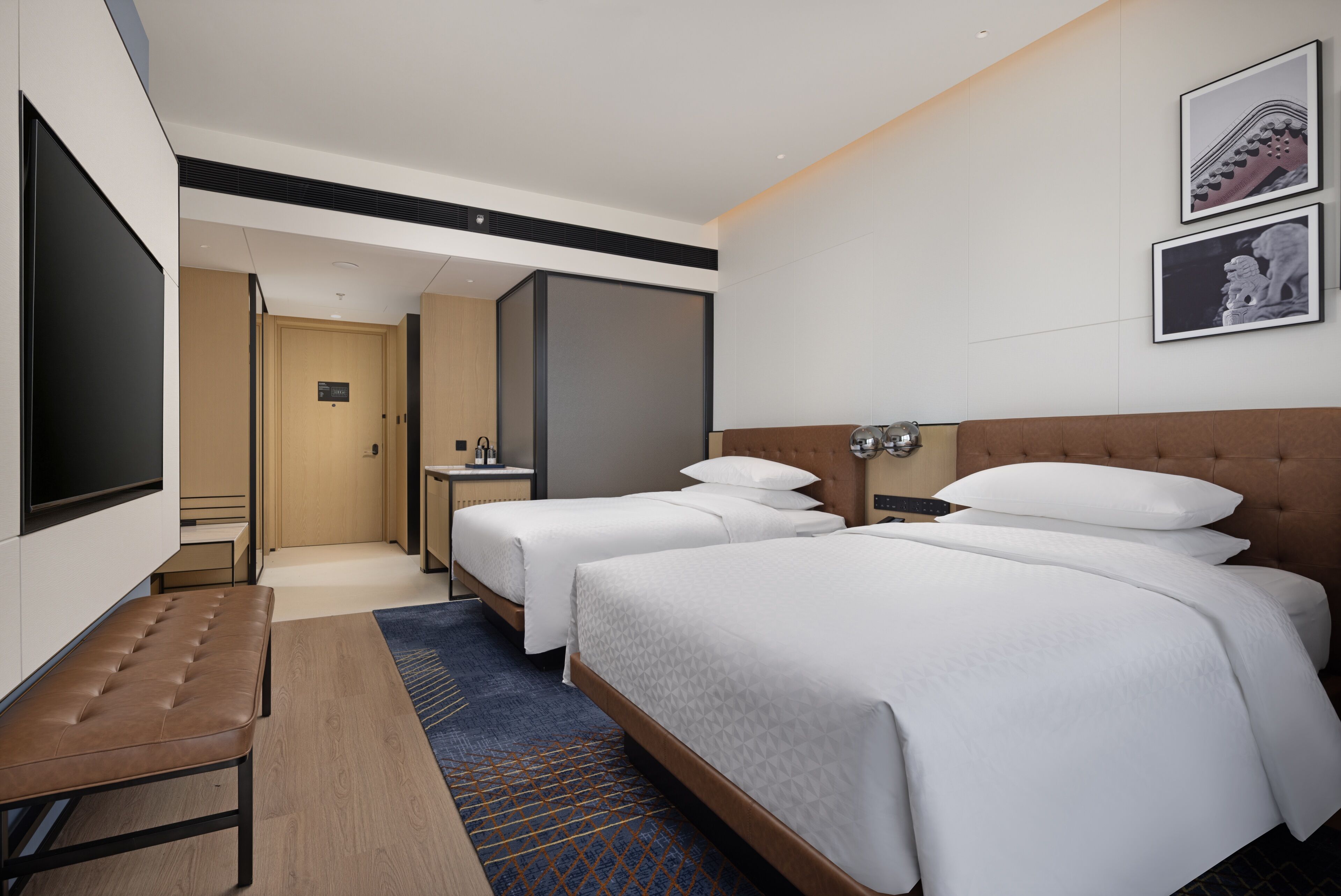 Une chambre d’hôtel avec deux lits, un téléviseur à écran plat, un banc, une petite table et une salle de bain visible à travers une porte ouverte.