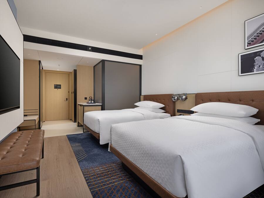 Une chambre d’hôtel avec deux lits, un téléviseur à écran plat, un banc, une petite table et une salle de bain visible à travers une porte ouverte.