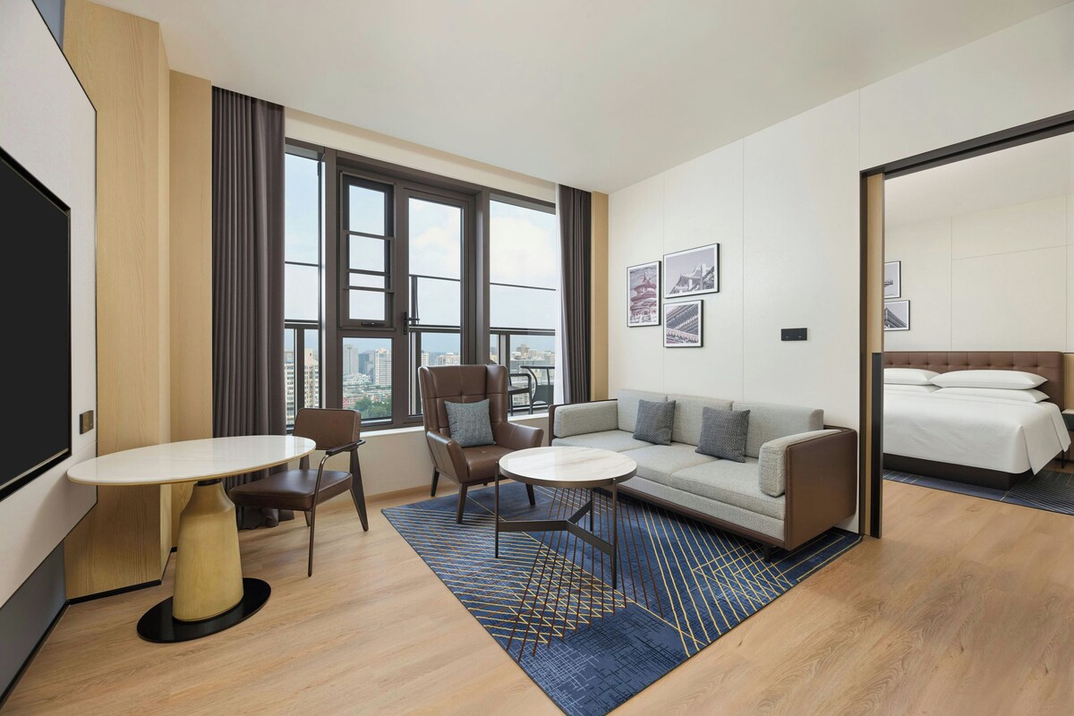 A hotel room with a sofa, a small table, a chair, a bed, and large windows offering a city view.