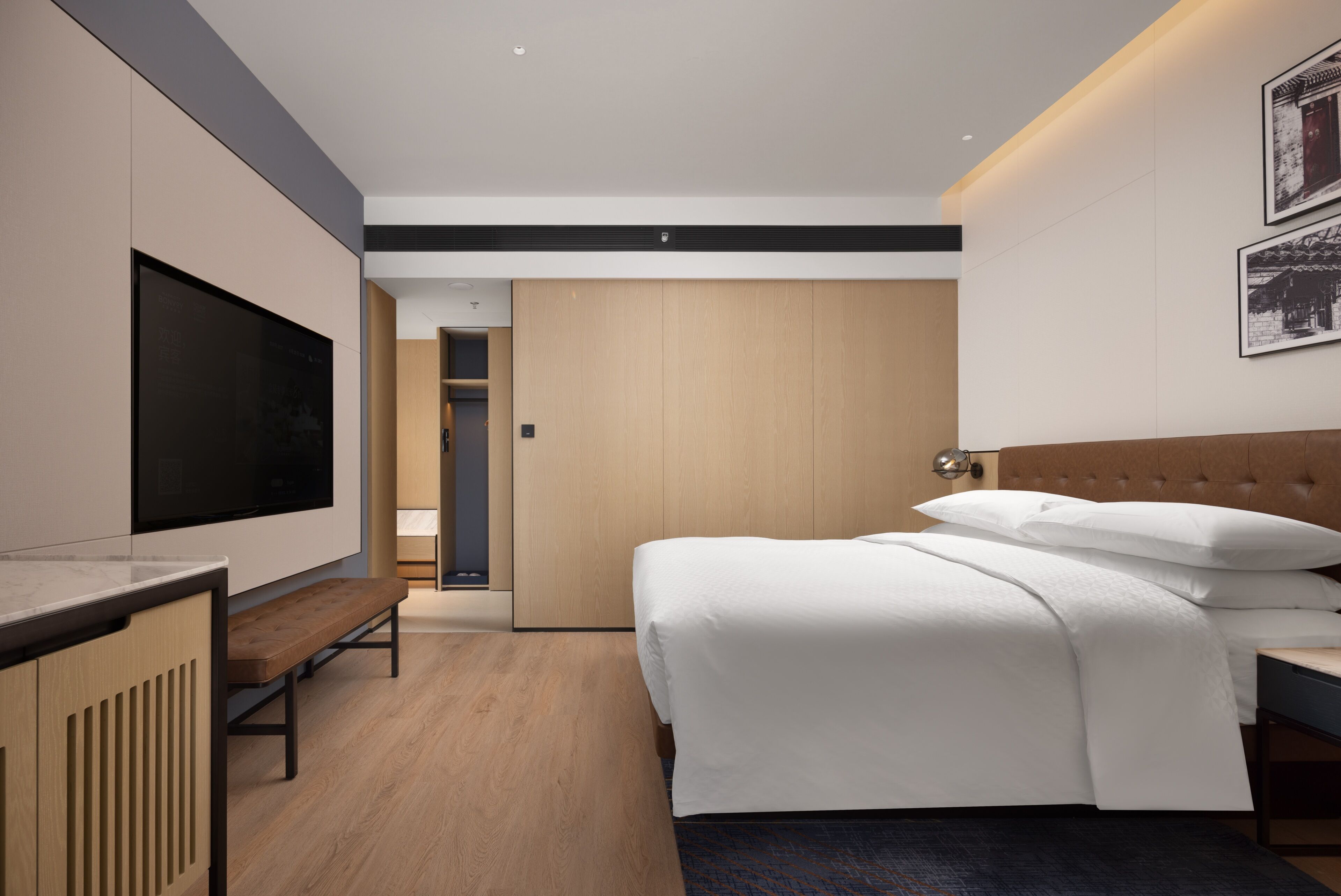 A modern hotel room with a large flat-screen TV, a wooden bench, a bed with white bedding, and a door leading to a bathroom.