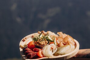 Una tabla de madera con diversos alimentos, incluyendo embutidos, verduras y un recipiente con salsa.