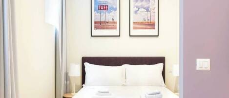Una habitación de hotel con una cama, dos cuadros en la pared, una mesita de noche y una lámpara.