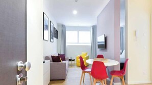Ein modernes Wohnzimmer mit einer Couch, einem runden Tisch und roten Stühlen. Ein Fernseher ist an der Wand befestigt und es gibt ein Fenster mit Vorhängen.