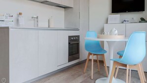 Eine moderne Küche mit weißen Schränken, einem Edelstahlbackofen und einem Flachbildfernseher, der über einem weißen Esstisch mit zwei blauen Stühlen angebracht ist.