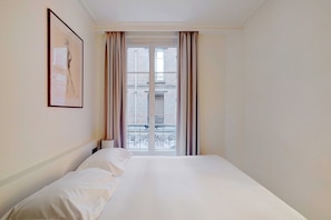 A hotel room with a bed, a framed picture on the wall, and a view of a building through the window. - Cozy Appartement a/c 1BR/2P- Montmartre (Paris)