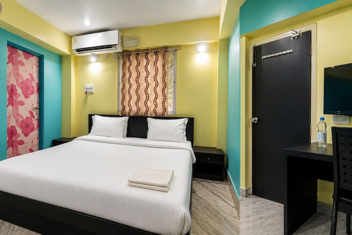 A hotel room with a bed, an air conditioning unit, a desk with a chair, and a door.